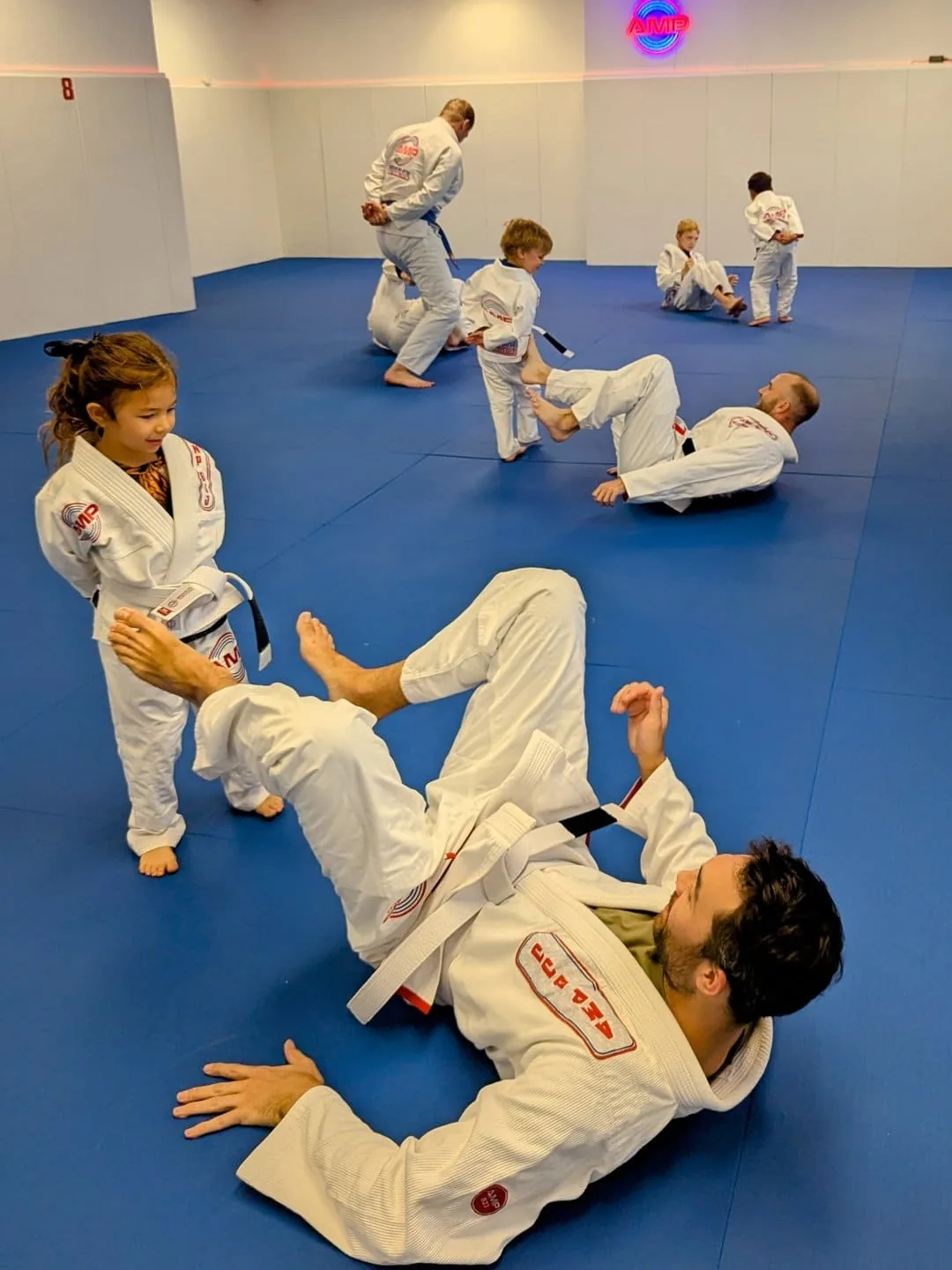 AMP BJJ family class in action — a young girl in a white gi stands beside her father drilling on the blue mats while other parents and kids train together in Austin, TX