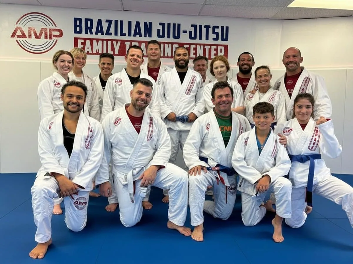 Group photo of AMP BJJ teens and adults in white gis smiling together in front of the Brazilian Jiu-Jitsu Family Training Center wall in Austin, TX